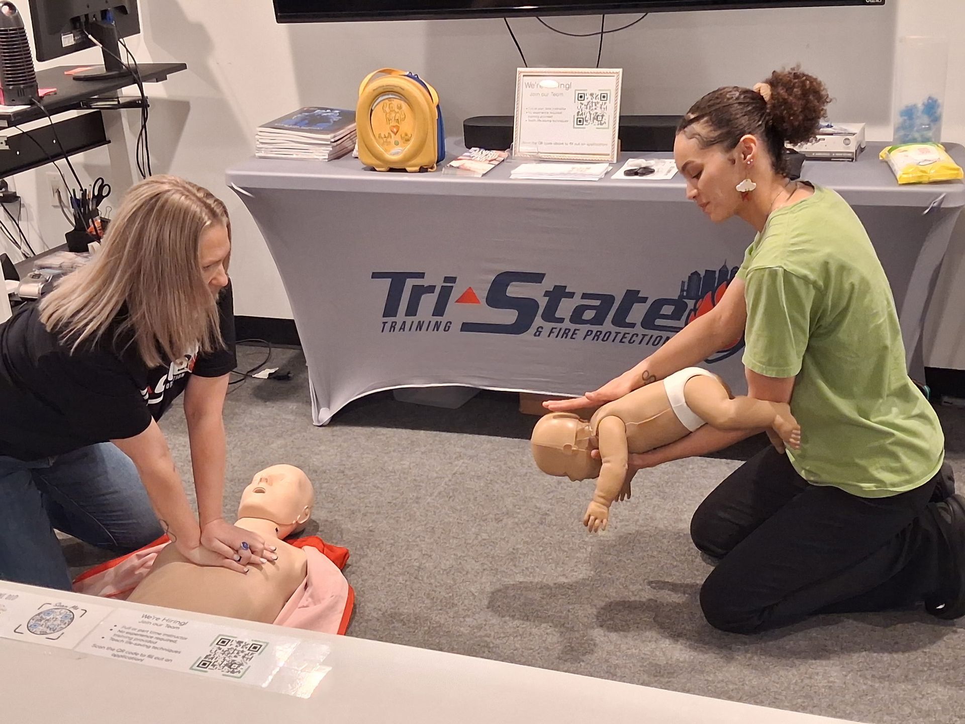 CPR training demo with instructor and participant practicing on a baby mannequin beside a Tri-State Training table
