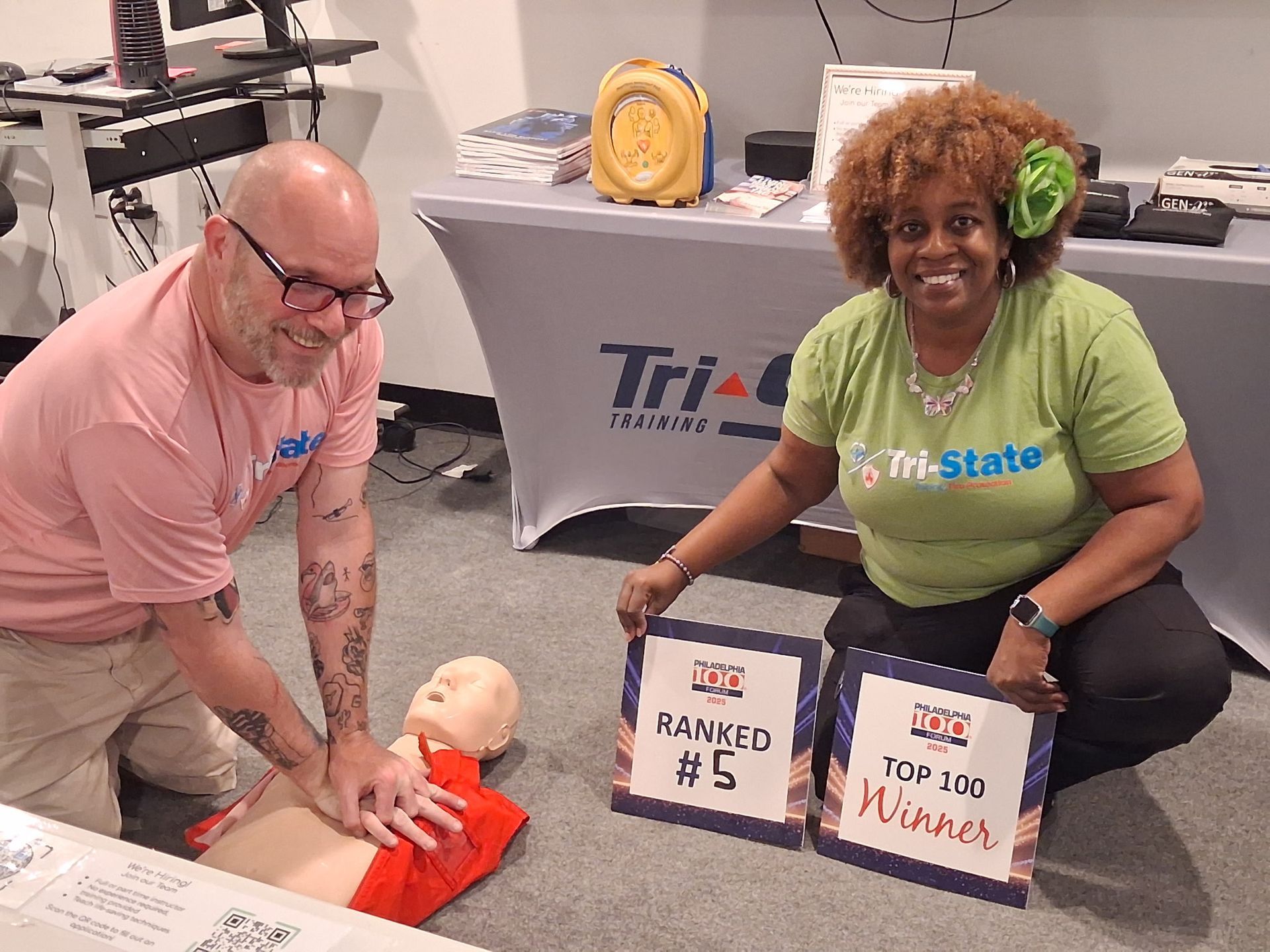 Two Tri-State Training instructors kneel beside a CPR manikin at a training booth, holding signs and smiling.