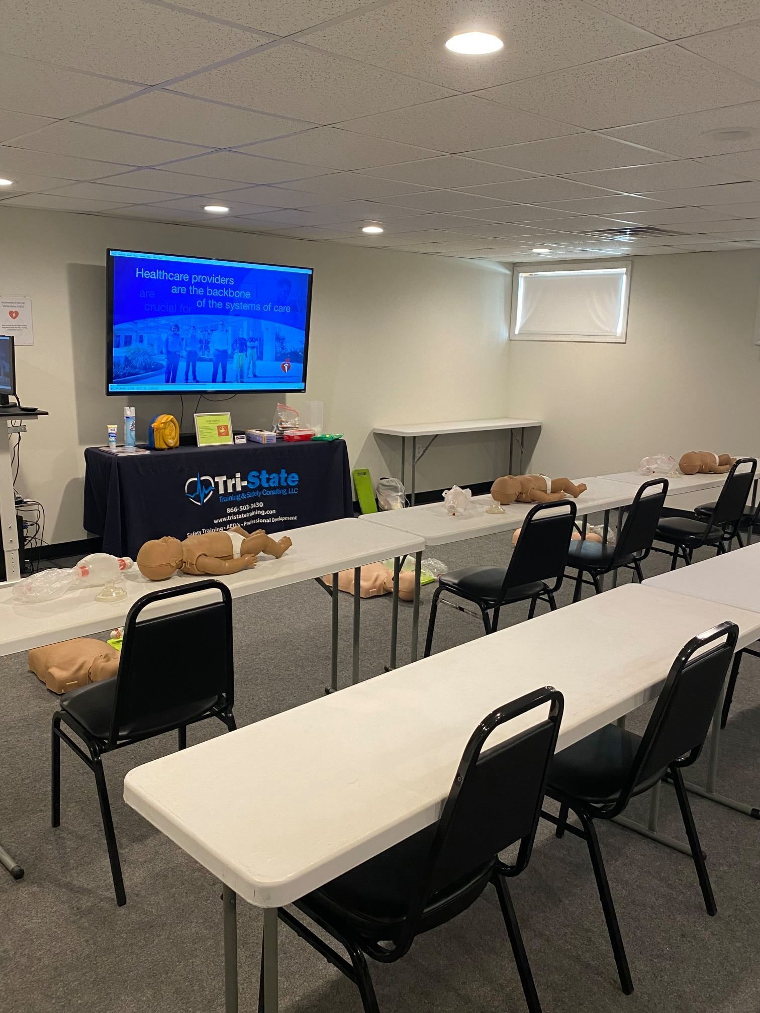 Empty conference room with long tables, black chairs, a projector screen, and CPR manikins on the table.