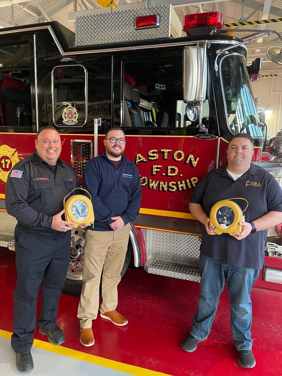 Three men in black and navy stand by a red fire truck holding an AED from Tri-State Training