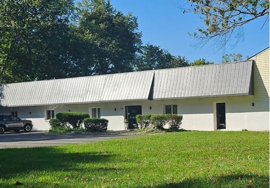 White single-story building with metal roof beside a grassy lawn and trees under a blue sky