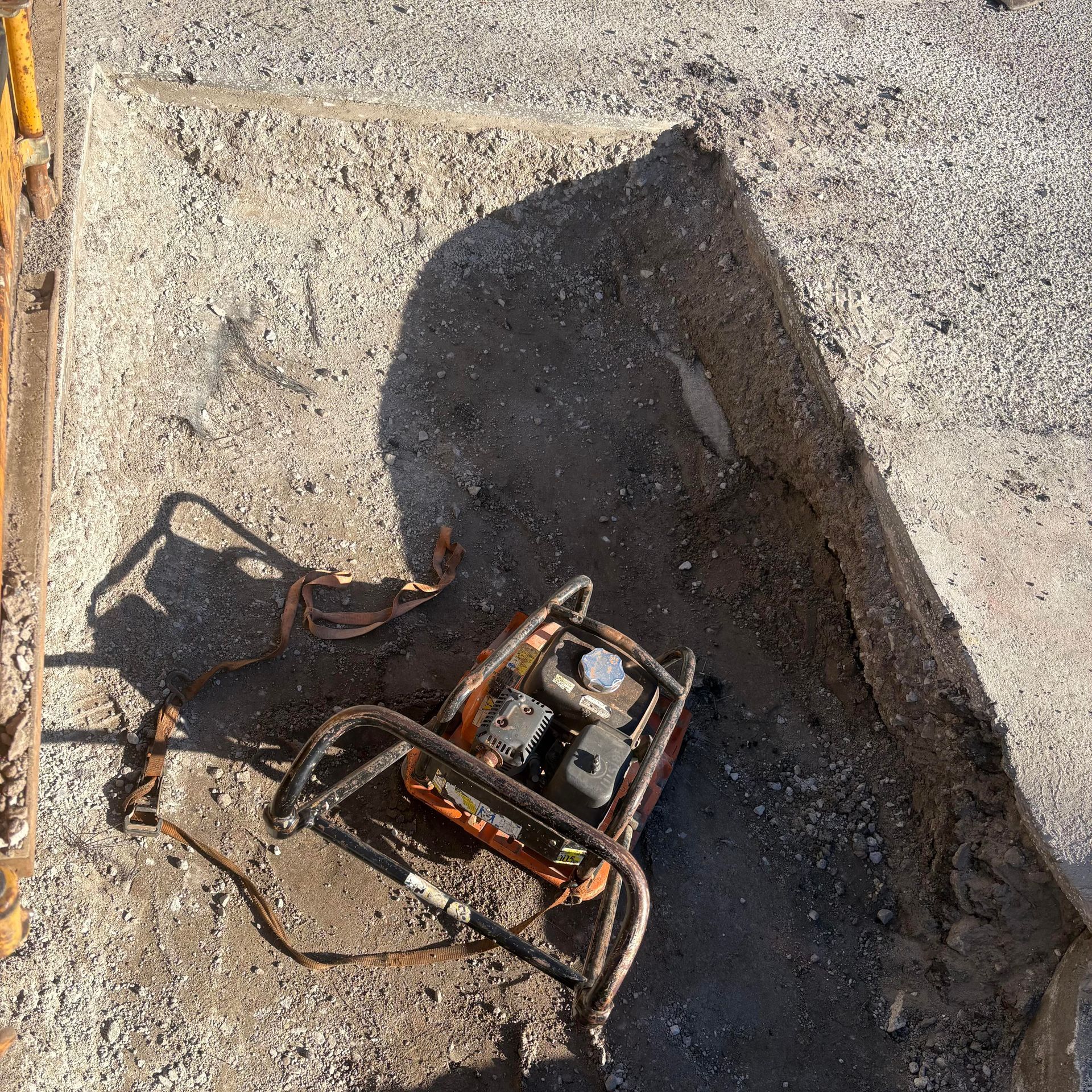 Rusty metal machinery in a dirt trench beside pavement, with shadows cast across the ground.