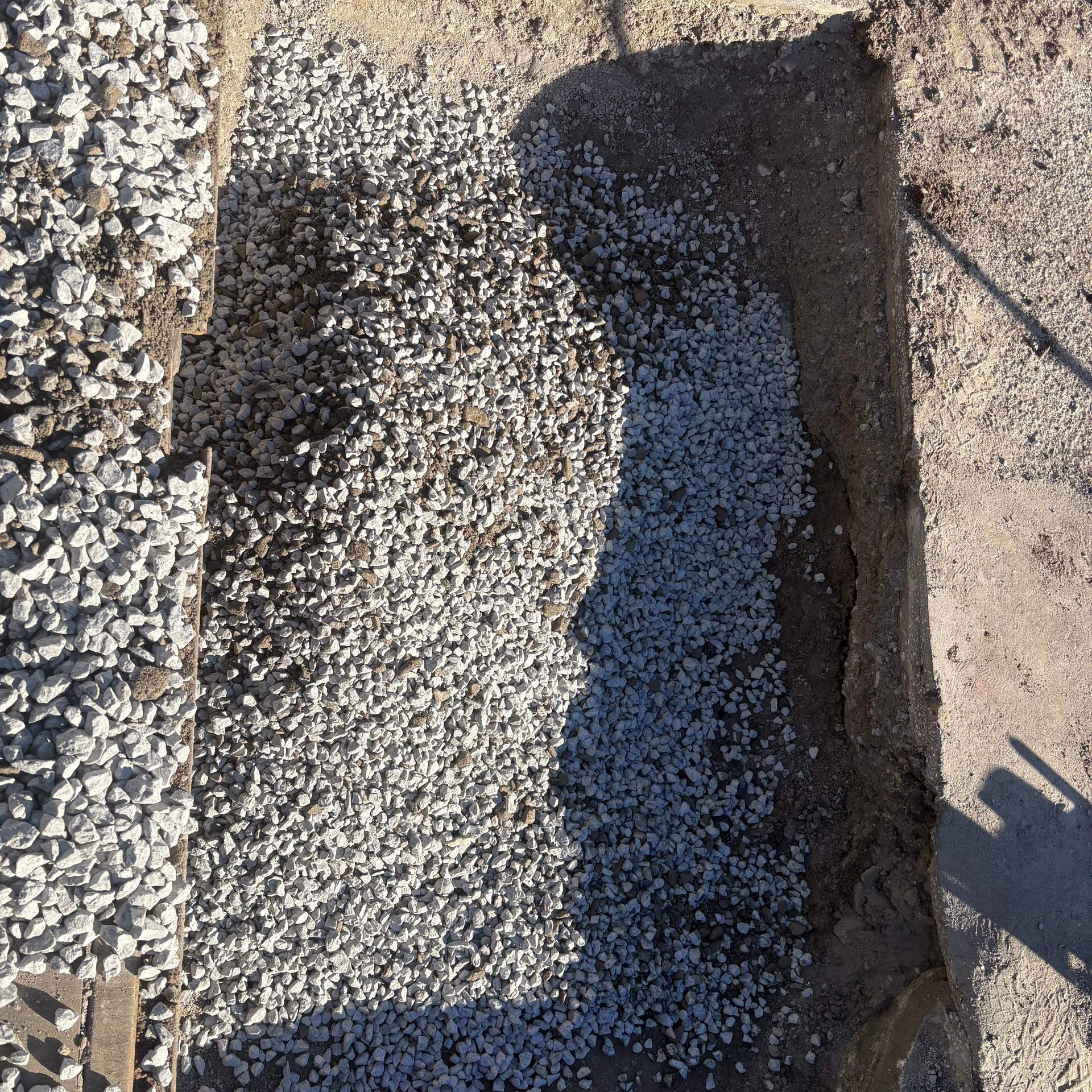 Gravel-filled rectangular trench beside a concrete edge, with shadow falling across the center