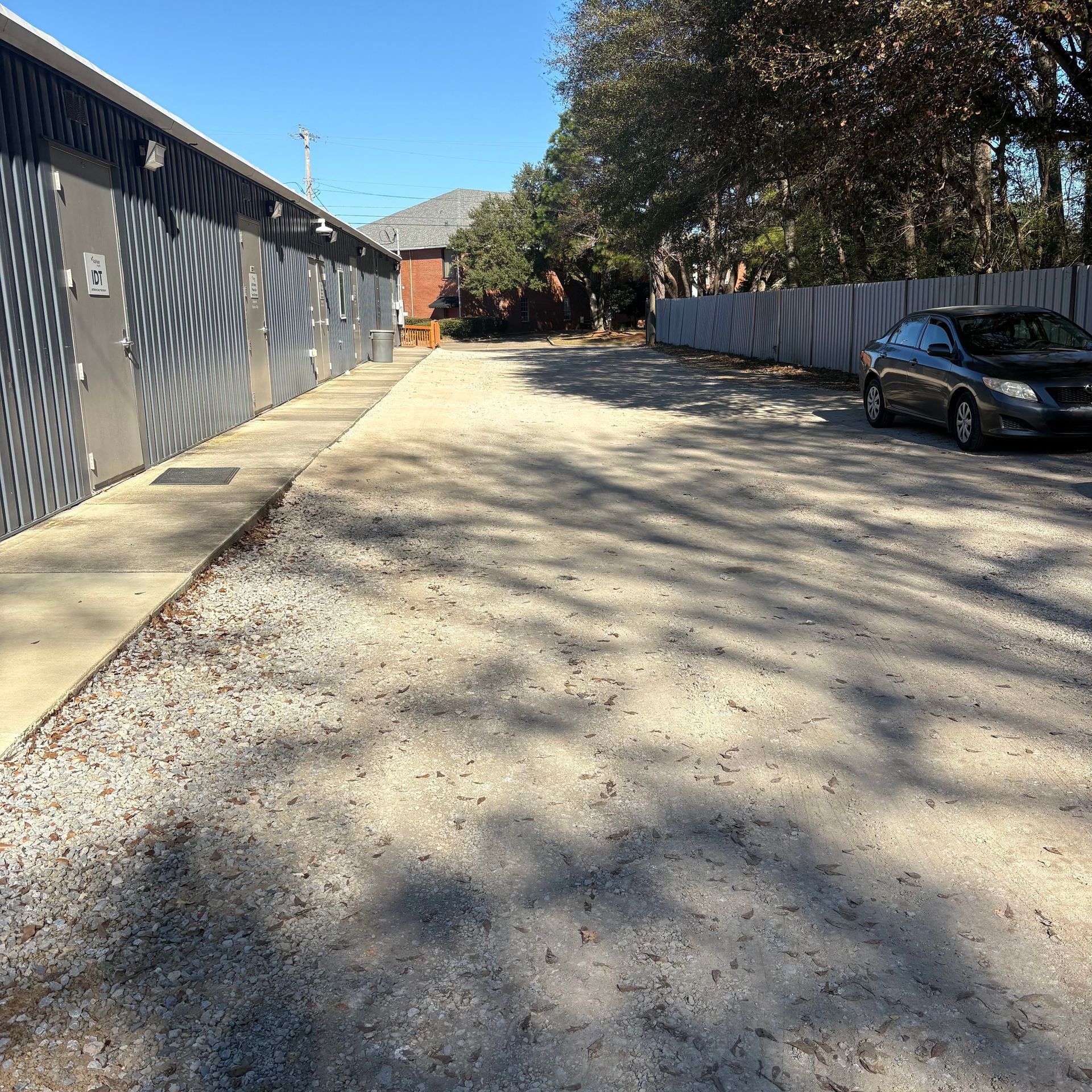 Sunny gravel driveway beside garages and trees, with a parked black car and shadows across the ground