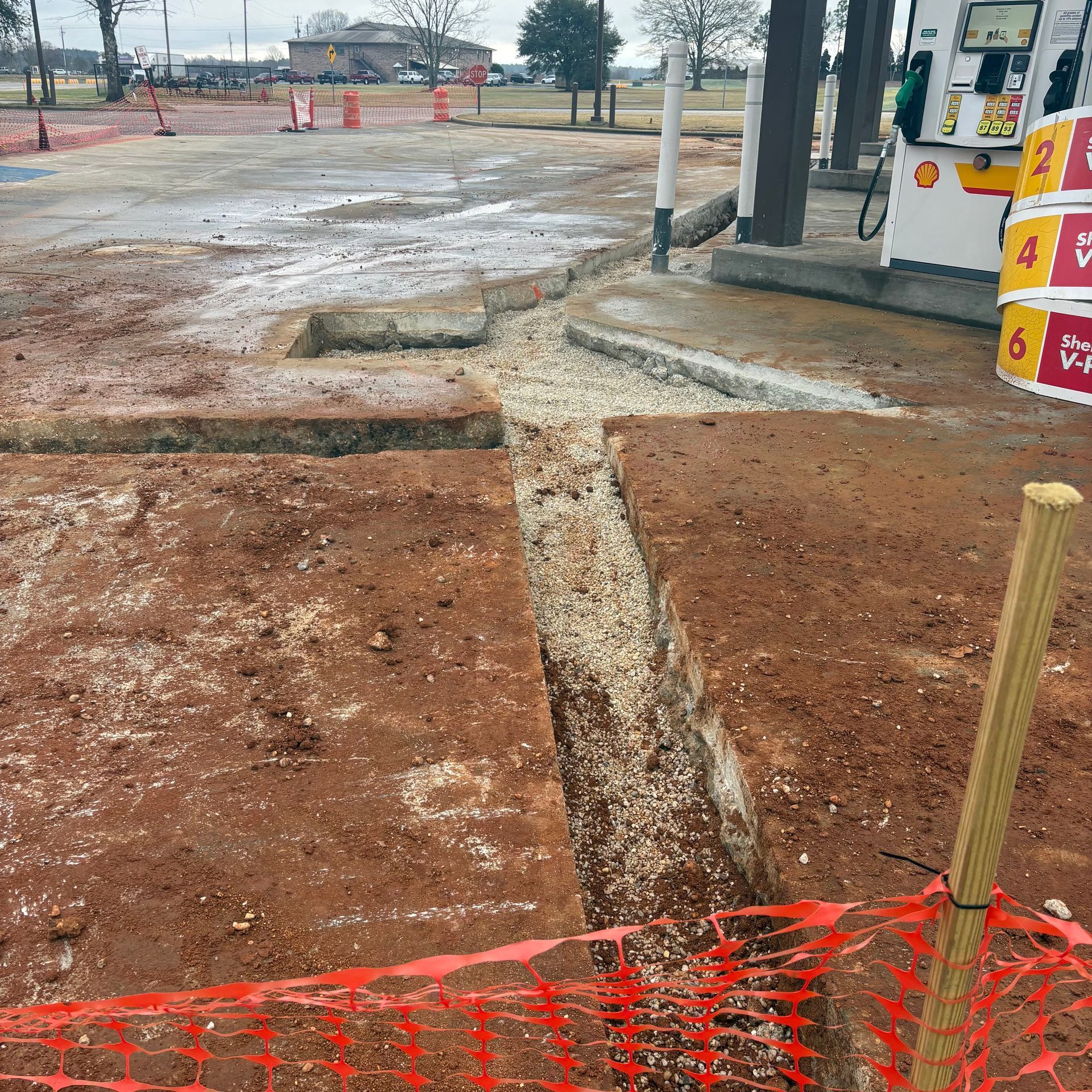 Construction site with muddy ground, trench lines, and orange safety fencing near a building entrance