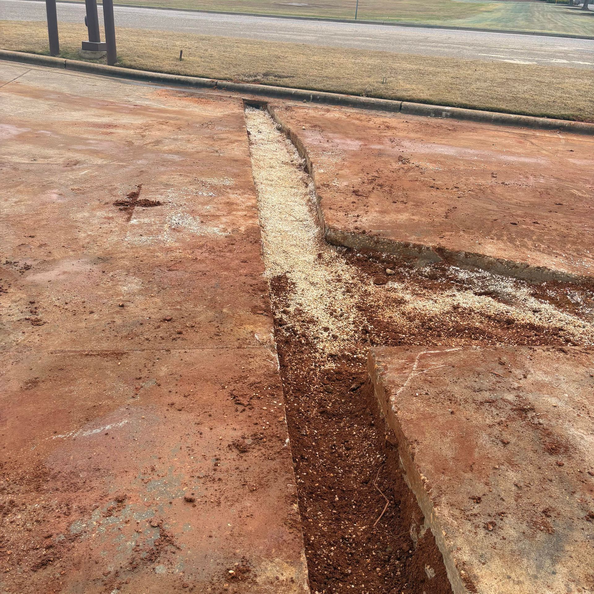 Cracked reddish concrete pavement with a long narrow trench of lighter gravel or soil running through it