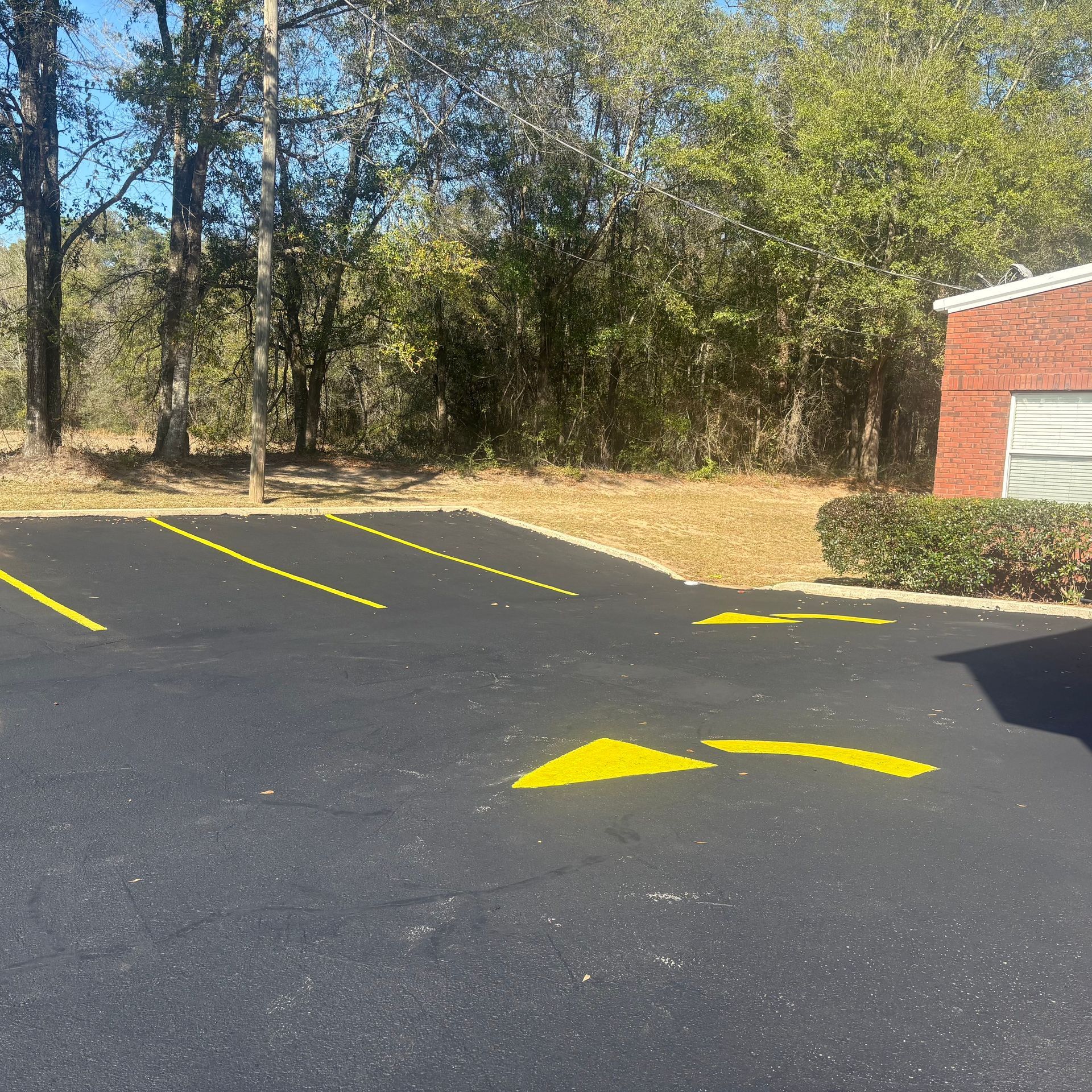 Empty parking lot with yellow arrows and diagonal lines beside trees and a brick building