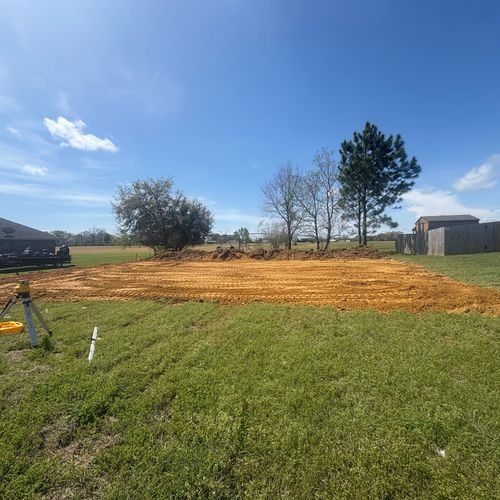 Sunny grassy yard with a freshly graded dirt patch, trees, and a small shed under a blue sky.