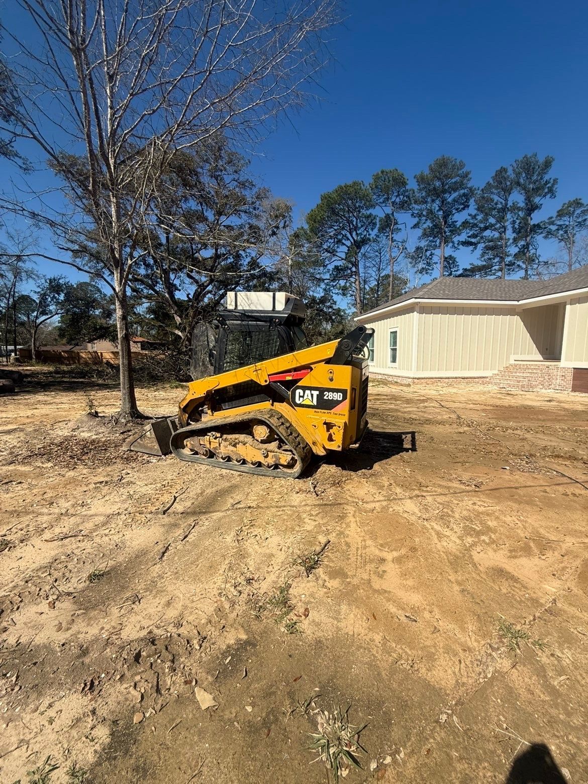 Yellow compact loader parked on a dirt lot beside a white house and bare tree under a clear blue sky