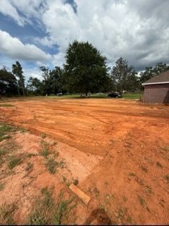 Dirt lot with orange soil, trees, cloudy sky, and a brick building on the right.