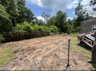 Cleared dirt lot beside trees and a building, with a parked white vehicle on the right.