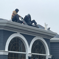 Two people crouch on a snowy rooftop above arched windows, wearing dark winter clothes.
