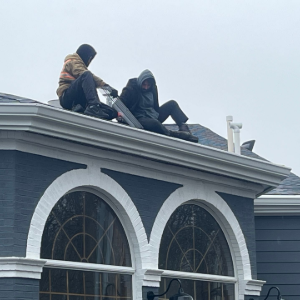 Two people crouch on a snowy rooftop above arched windows, wearing dark winter clothes.