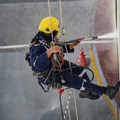 Rope-access worker in yellow helmet rappels beside a building, spraying water onto the wall.
