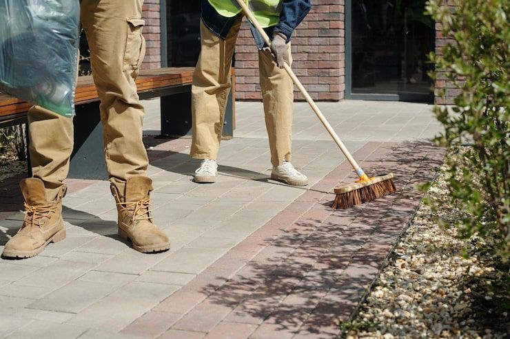 Workers sweeping a sidewalk beside a building, with brooms and tan work pants.