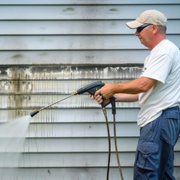 Man in cap pressure-washing gray house siding with a hose wand