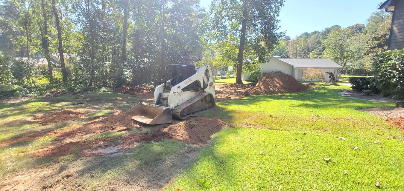 A white excavator and dirt pile in a wooded yard near a small shed on a sunny day