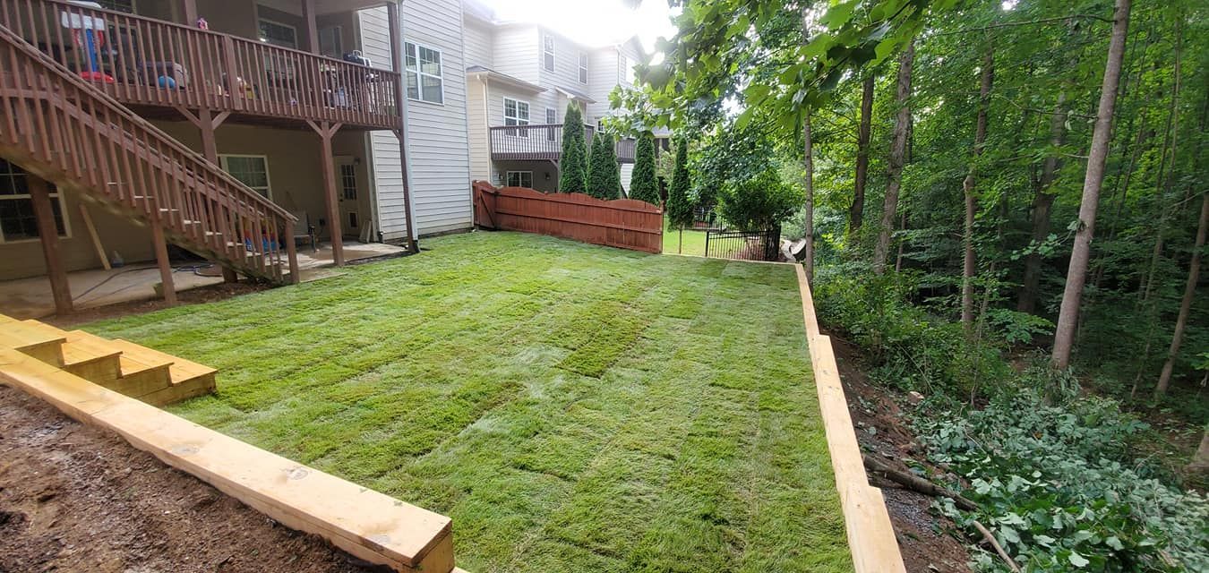 Backyard lawn beside a wooden deck, with trees and a narrow gravel path along the edge