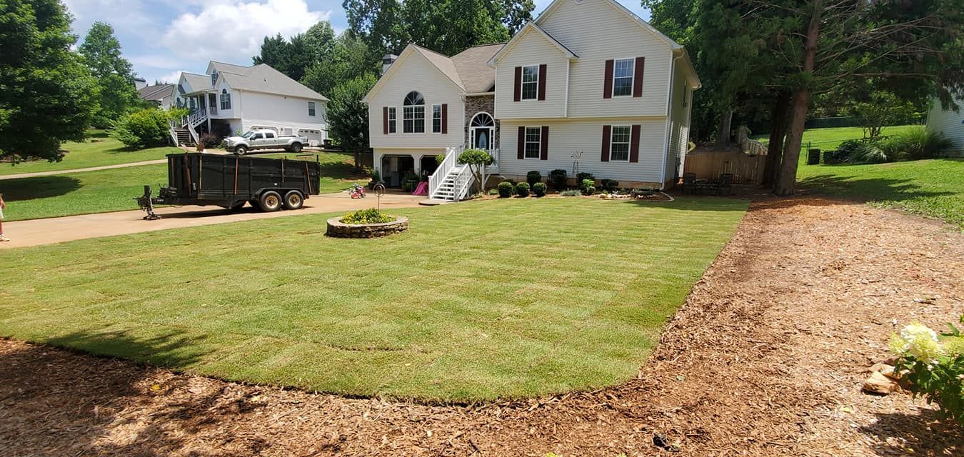 White house with black shutters and a truck in front, on a grassy yard with a mulch path and trees.