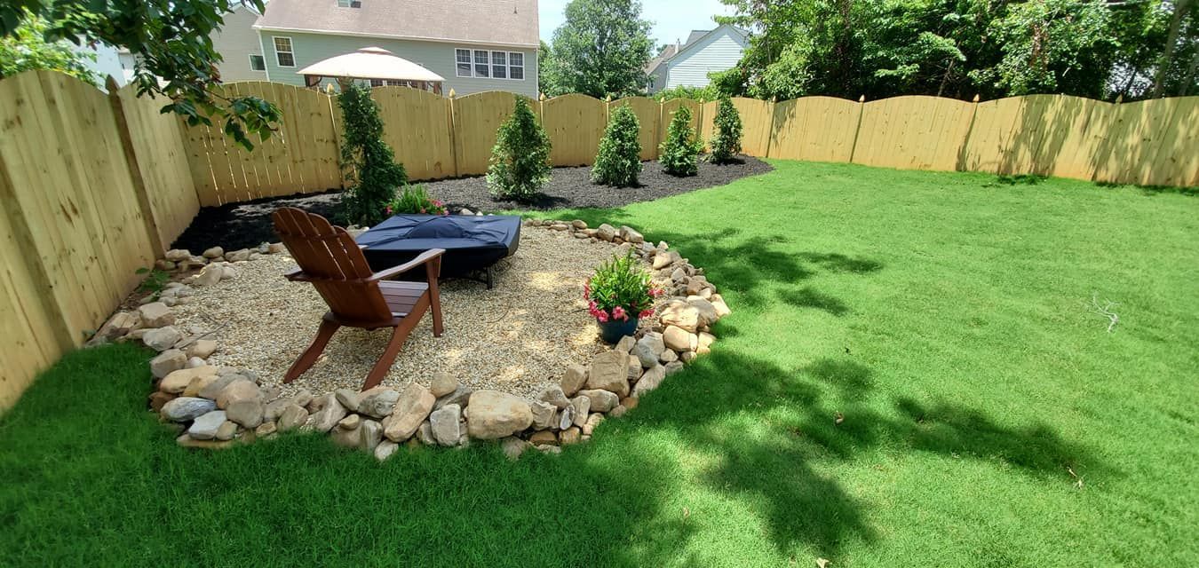 Backyard with a stone fire pit, Adirondack chairs, and green lawn bordered by a wooden fence.