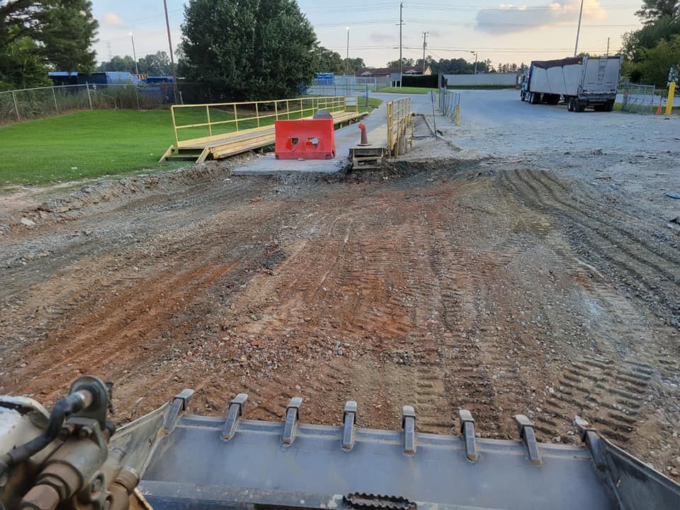 Construction site with gravel road, dirt piles, orange barriers, and a loader bucket in the foreground