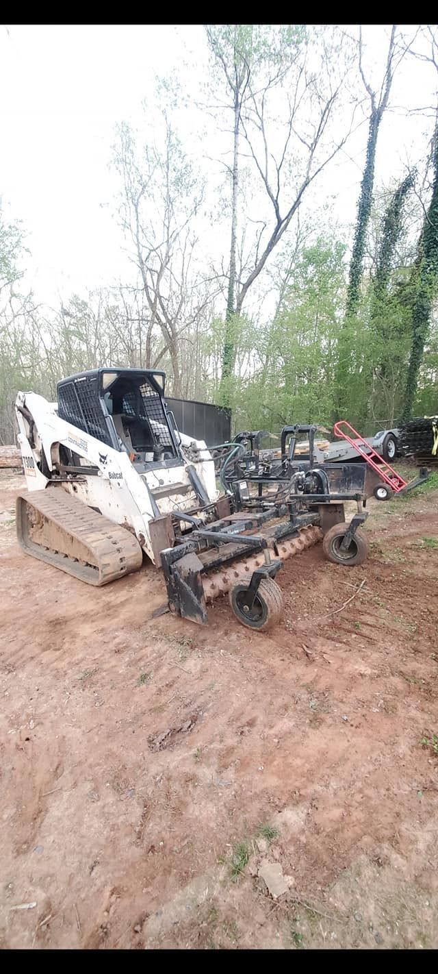 White skid-steer loader with damaged front attachment on muddy ground in a wooded area