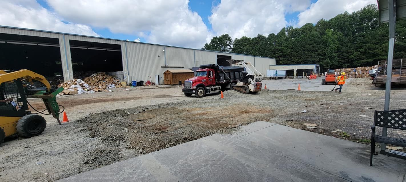 Construction yard with loader and dump truck near a warehouse, piles of gravel and debris under a partly cloudy sky