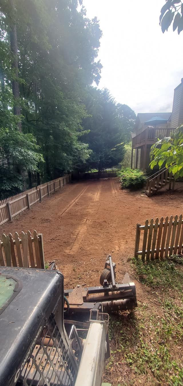 Wooded dirt path with a small fence and machinery in the foreground, leading toward houses in the distance
