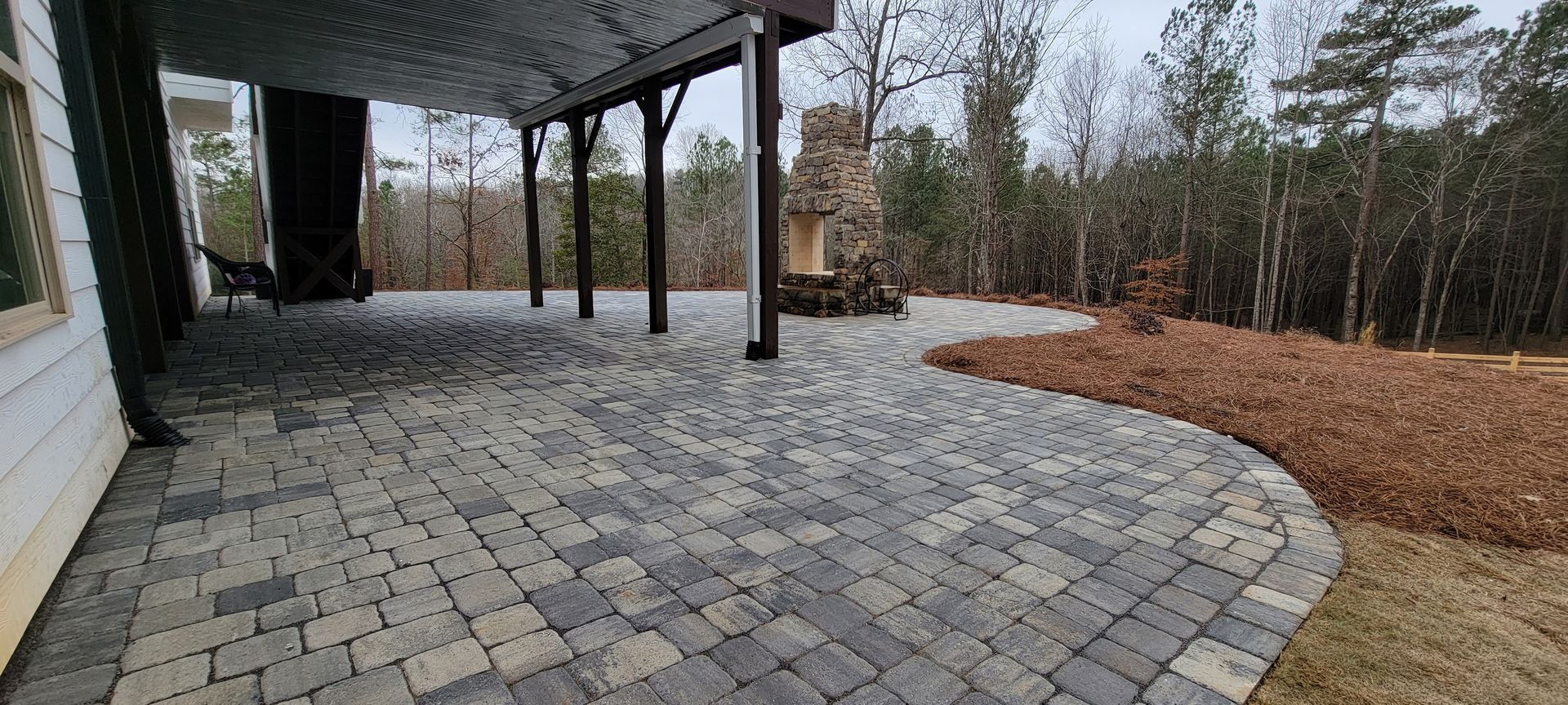 Covered patio with gray paver floor, black posts, and a curved mulch garden bed by trees