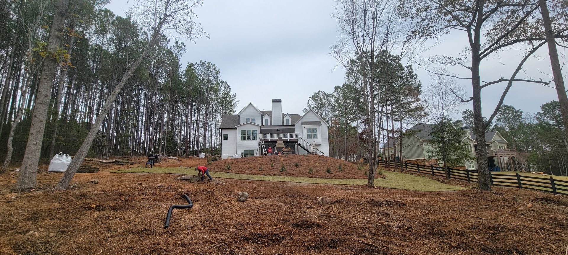 Large white house beyond a cleared wooded lot with bare trees and a wooden fence under a cloudy sky