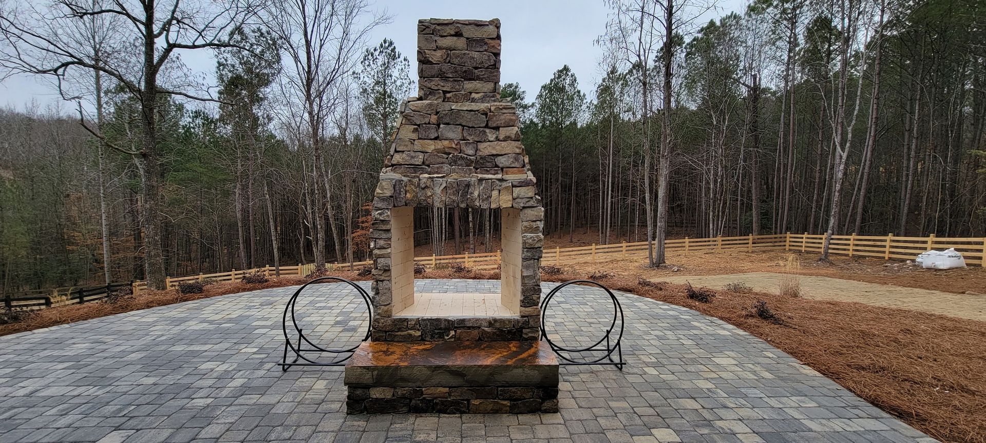 Stone chimney-style monument on a brick patio in a wooded park, with circular metal rings on both sides.