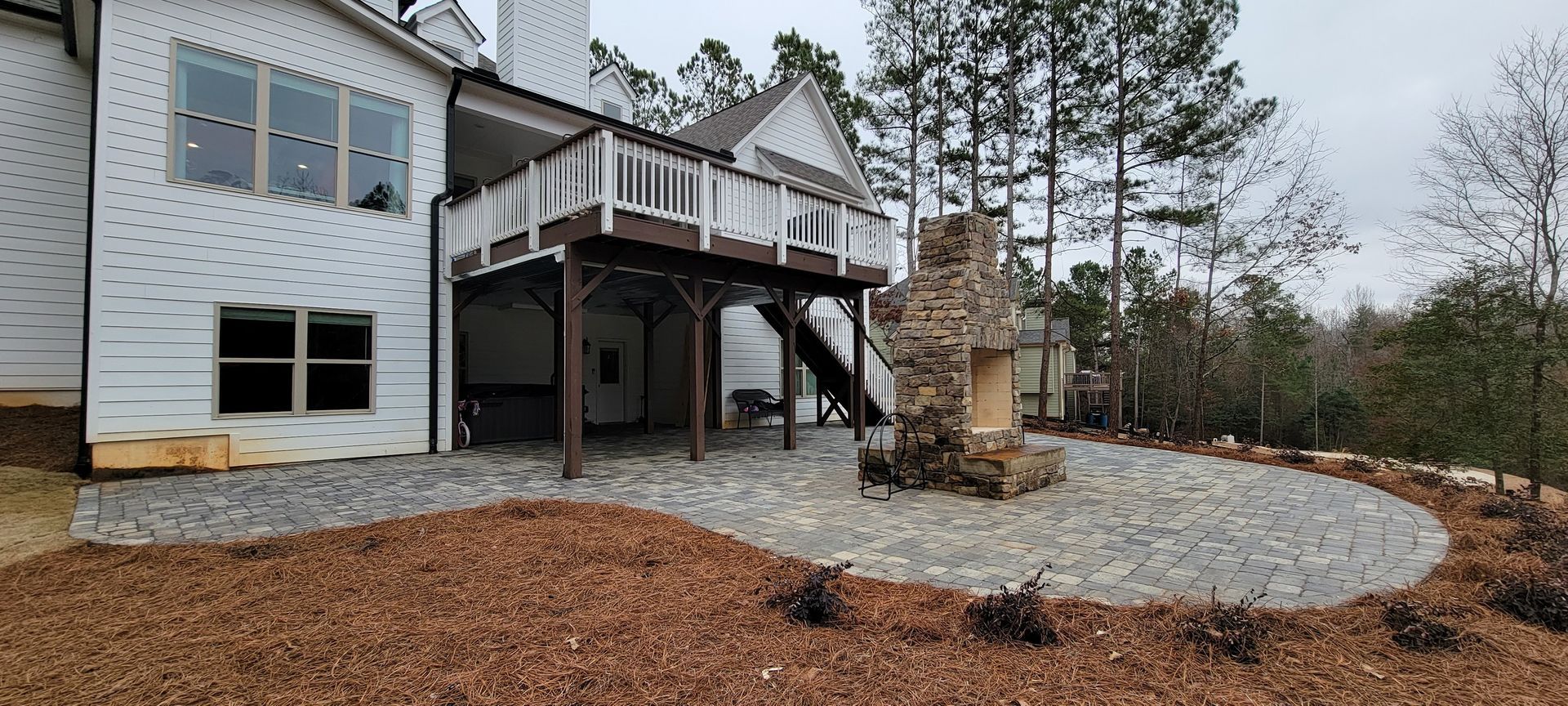 Two-story house with raised deck, stone fire pit, and wood-chip landscaping in a wooded yard