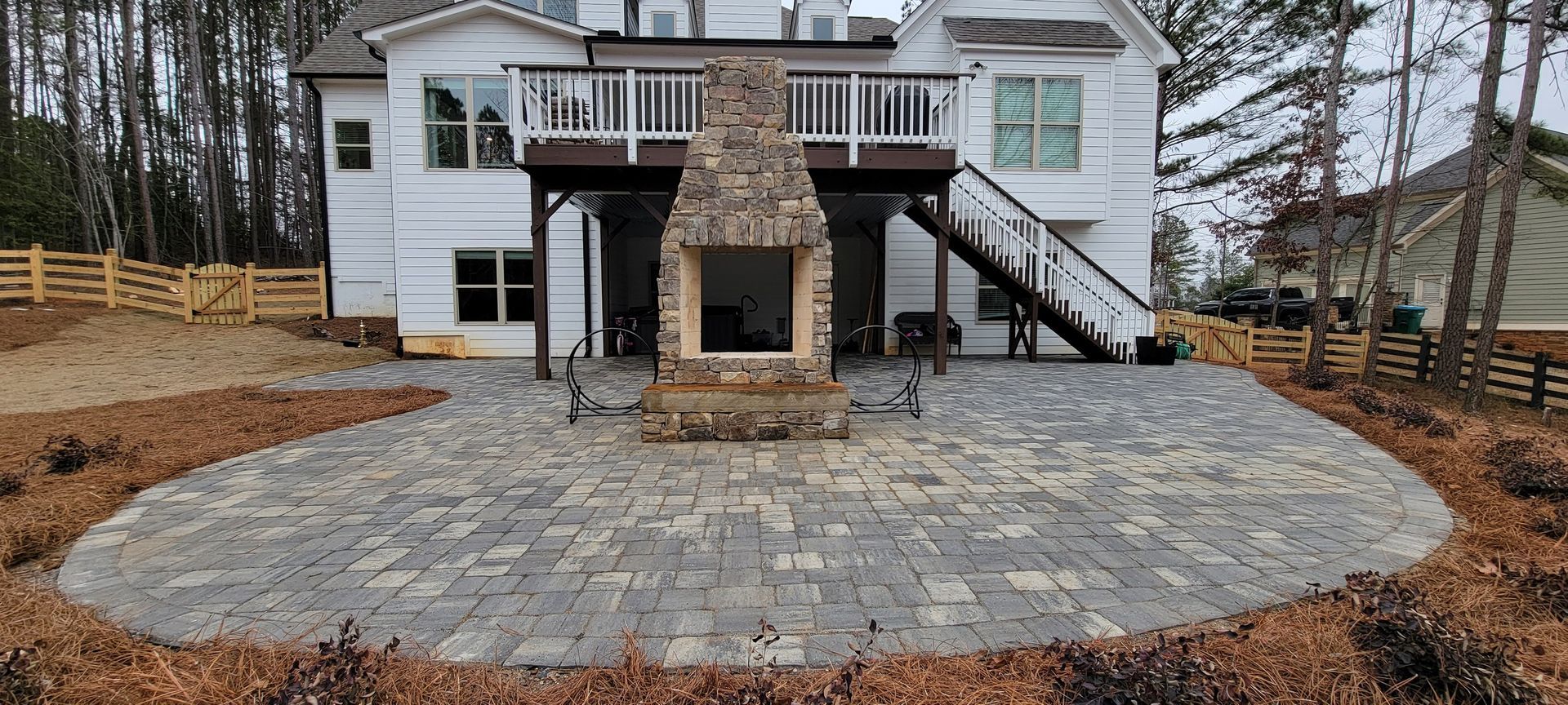White two-story house with stone outdoor fireplace and large paver patio in a wooded yard