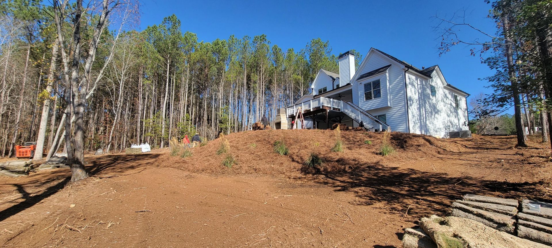 White two-story house on a cleared wooded hillside under a bright blue sky