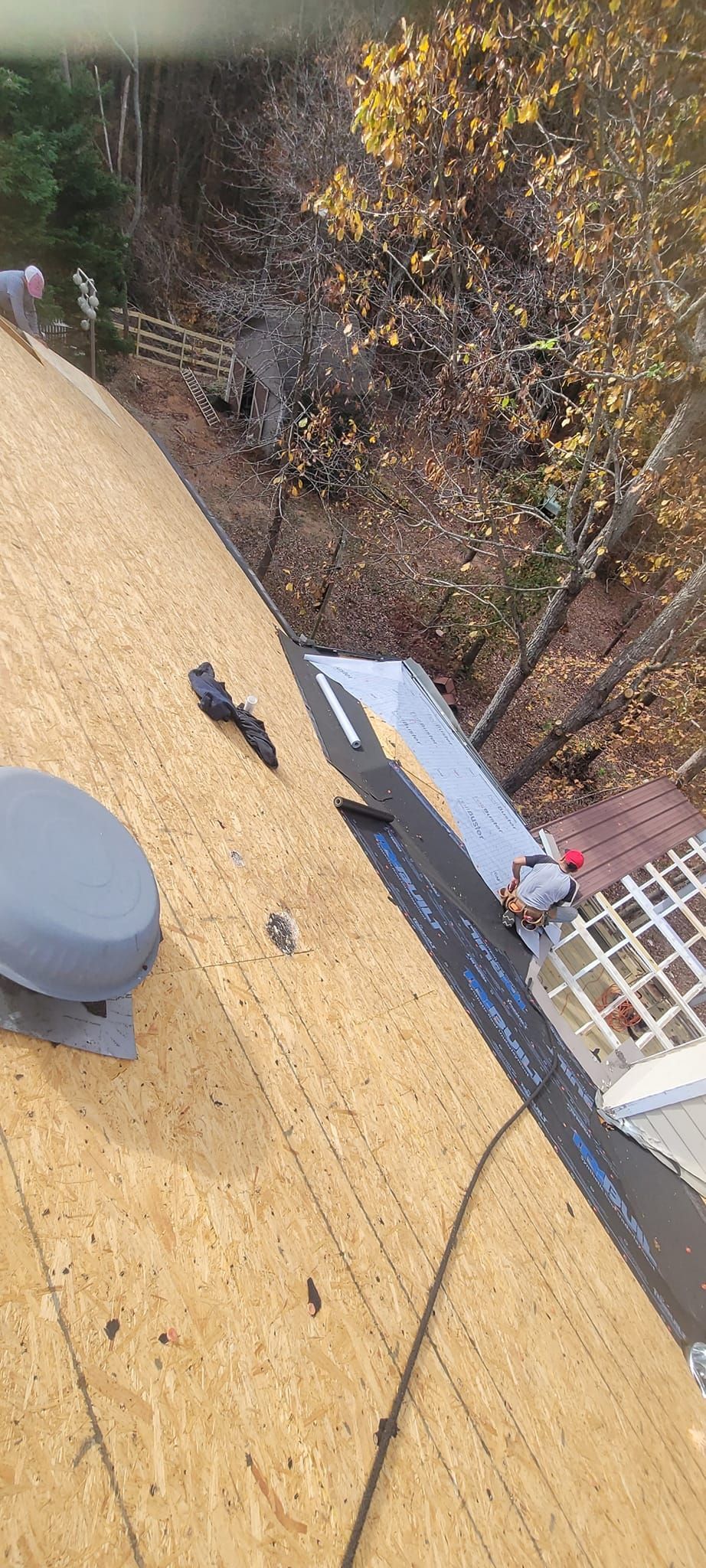 Tilted view of a roof with tan shingles, a vent, and autumn trees below near a house