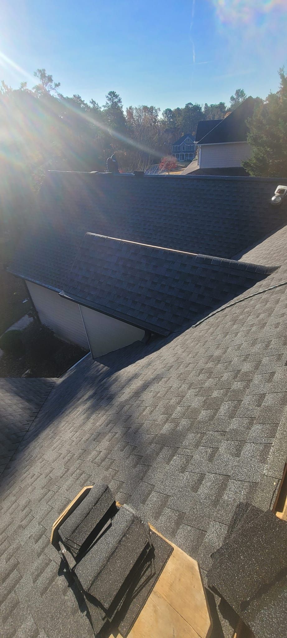 Rooftop with dark shingles under bright sunlight, trees and blue sky in the background.