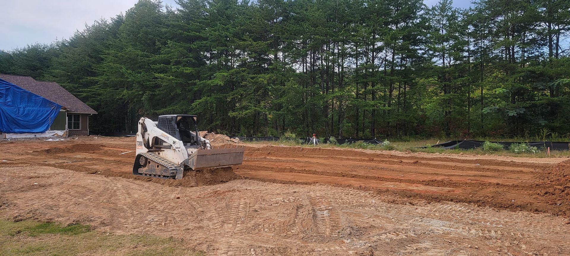 Small excavator grading a dirt lot beside a tree line and a tarp-covered structure.