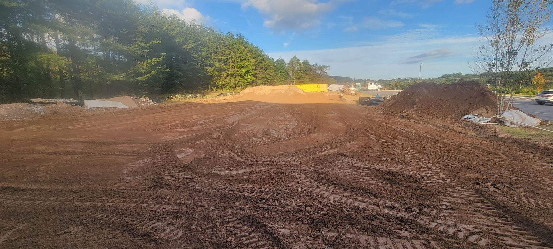 Wide dirt lot under construction with trees, a blue sky, and yellow equipment in the distance