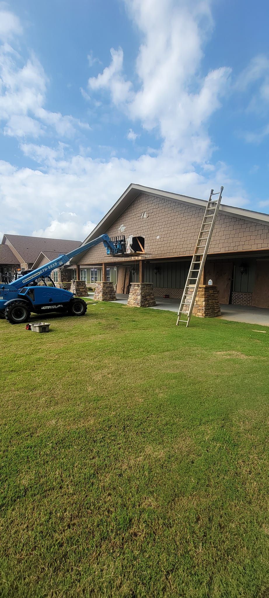 Blue cherry picker in grassy yard beside a tan house under a partly cloudy sky