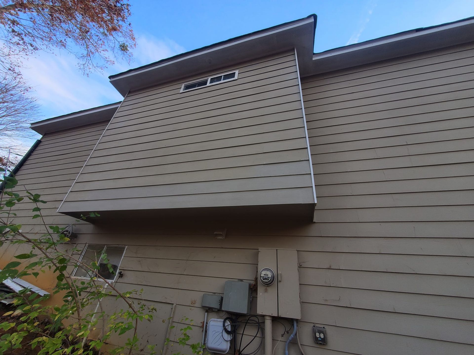 Exterior of beige siding house with a protruding second-story bump-out and utility meters below