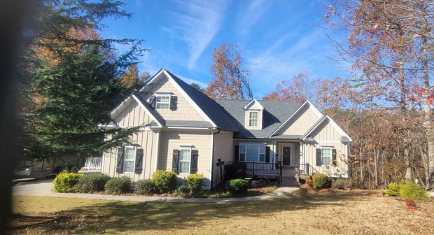 Suburban house with beige siding, dark roof, and front porch surrounded by trees in autumn.