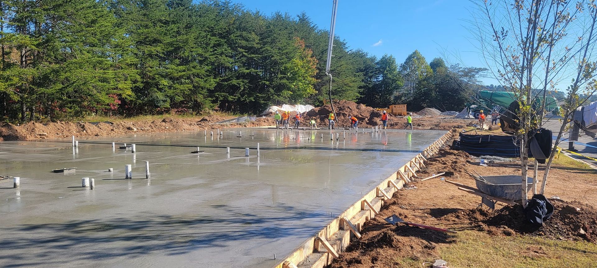 Concrete foundation under construction beside a dirt road and trees, with workers in the distance.