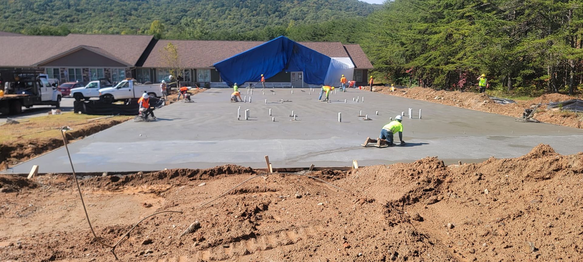 Workers pouring concrete foundation at a construction site with blue tarp-covered structure in the background
