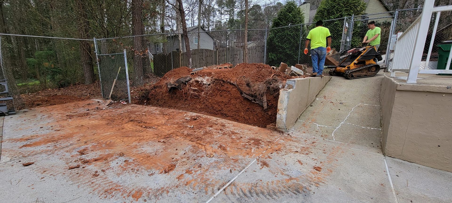 Workers inspect a large sinkhole beside a driveway, with excavated dirt and a damaged retaining wall.