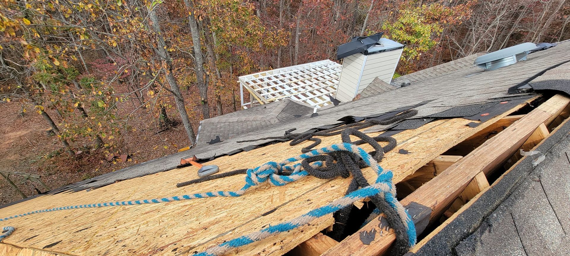 Roof damage with exposed plywood and blue tarp straps on a house in a wooded area