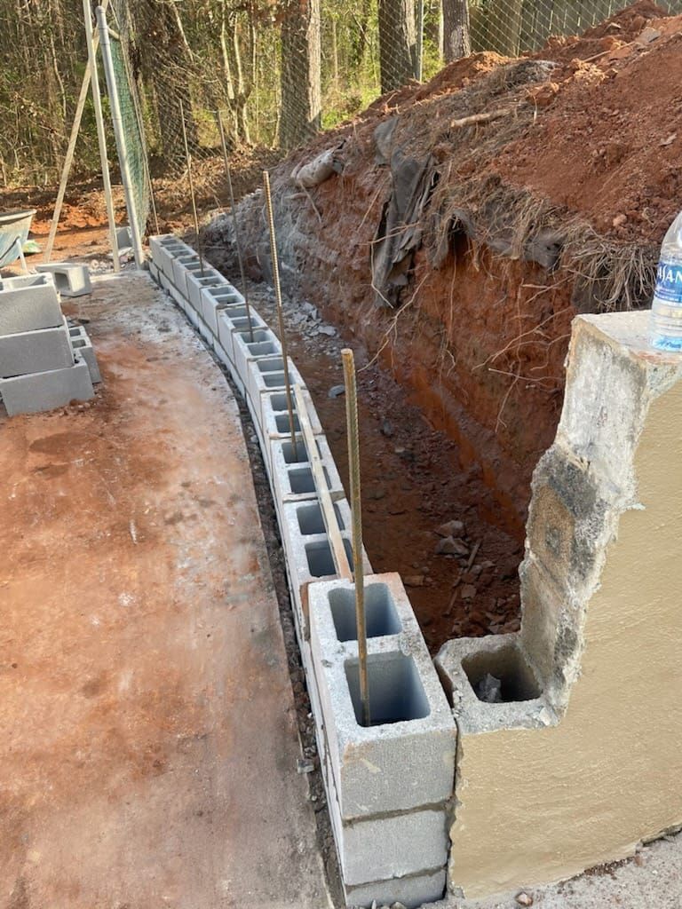 Curved cinder block retaining wall under construction beside a red dirt slope in a wooded area