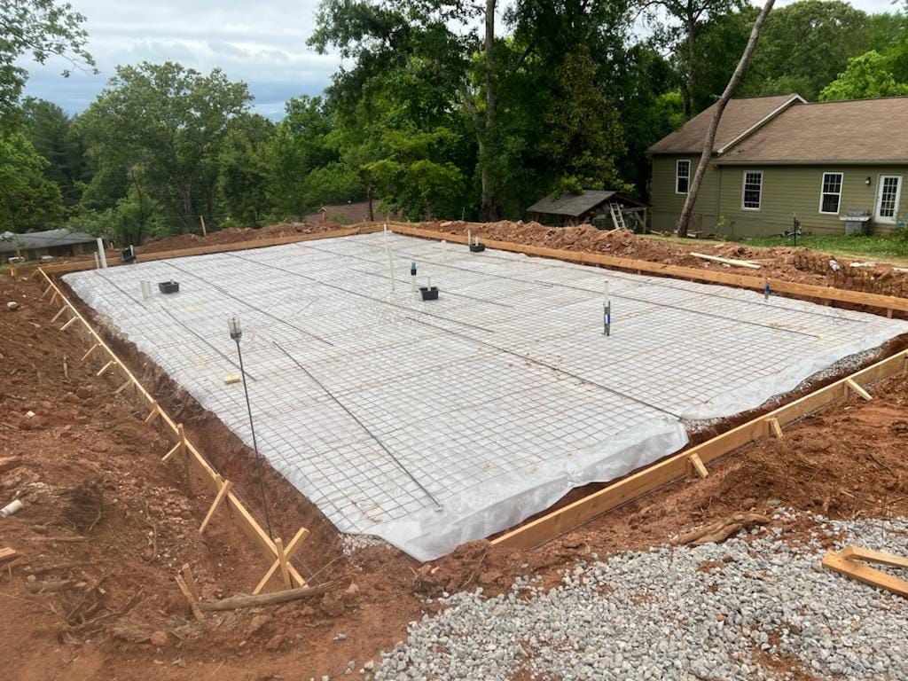Foundation slab with white vapor barrier and exposed rebar at a construction site near a house