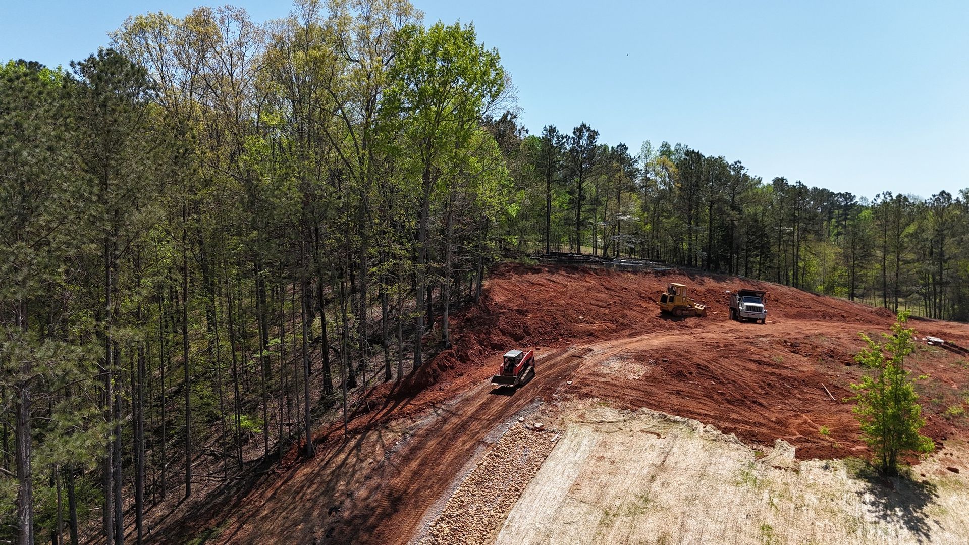 Bulldozers grading a red dirt clearing beside a wooded hillside under a blue sky