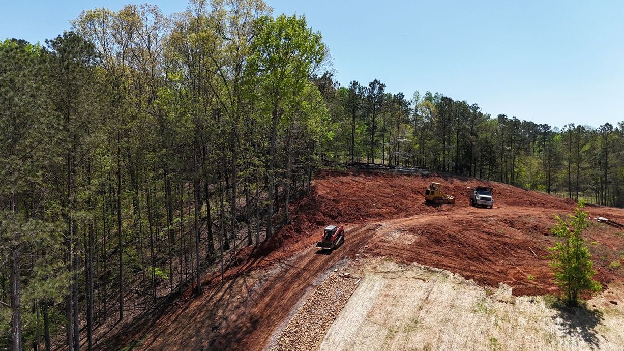 Bulldozers grading a red dirt hillside beside a wooded forest under a clear sky