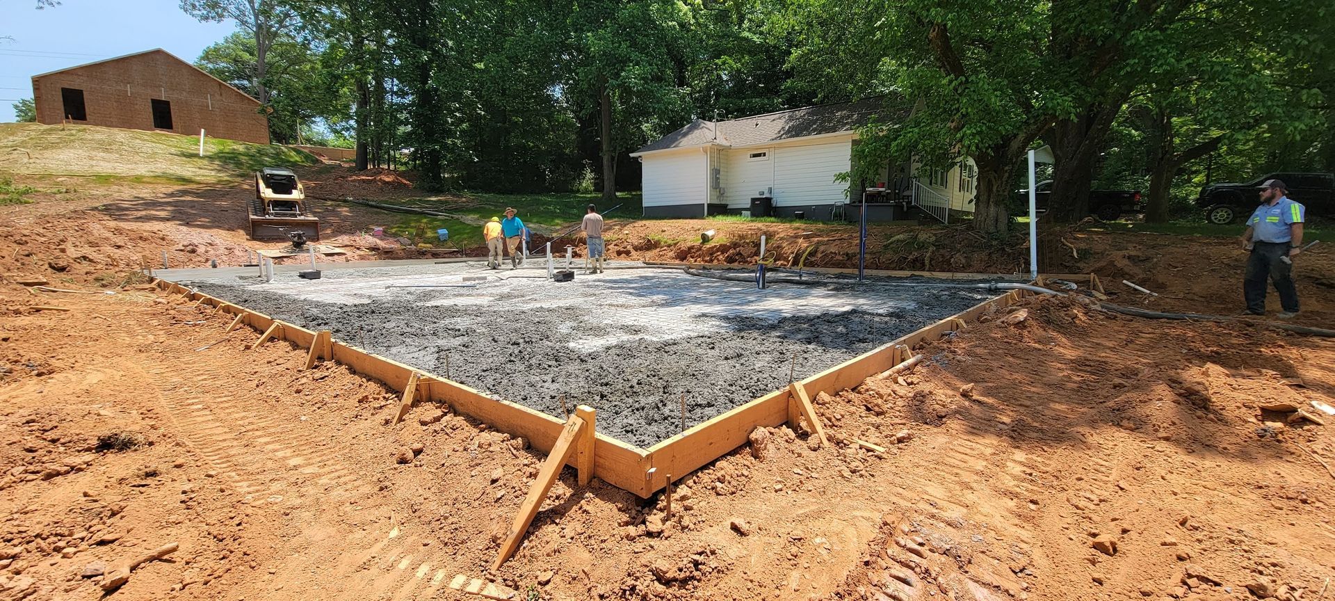 Foundation slab under construction in a dirt lot, with workers, trees, and a small building nearby.